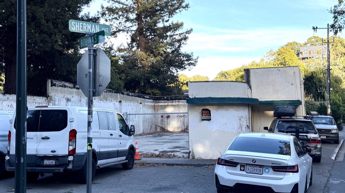 The fenced, cleared site of the burned dry cleaner at Bolinas Road and Sherman Avenue, with a surviving stub of wall.