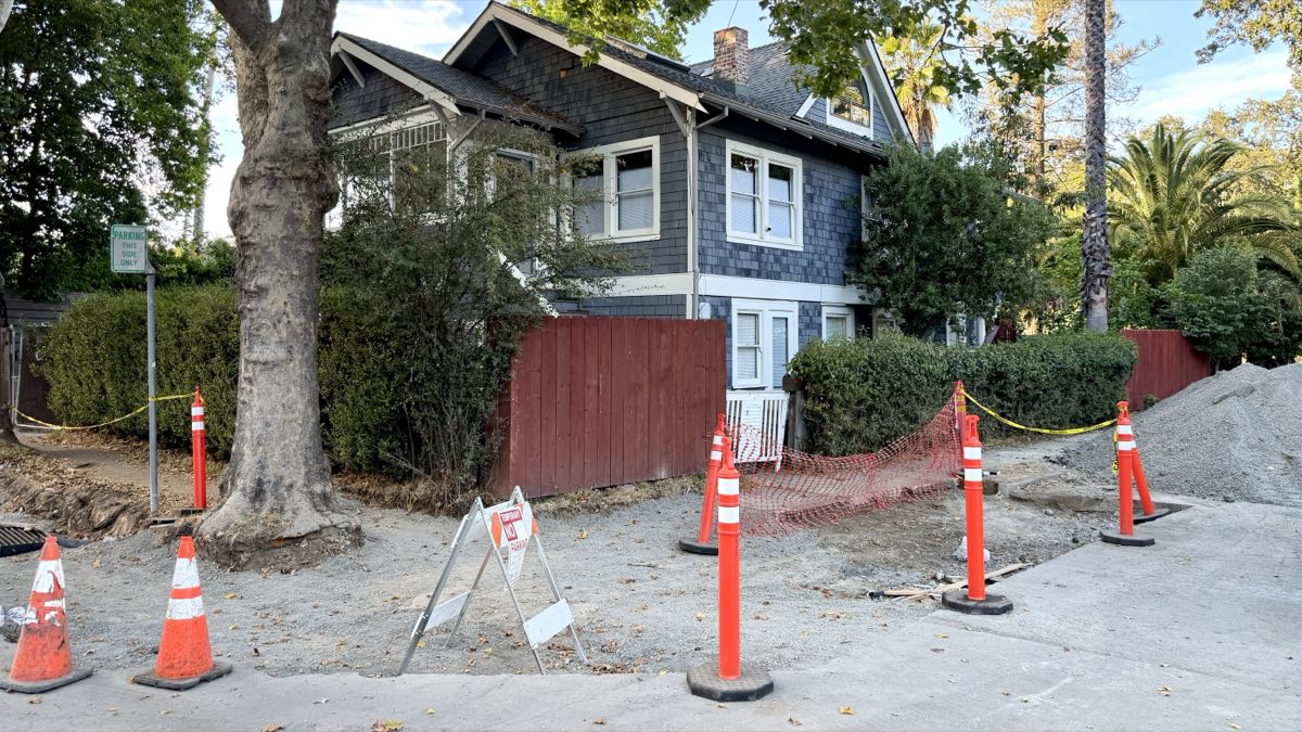 Traffic cones, orange safety netting and a gravel pile across a dug-up stretch of Fairfax pavement, with a shingled house behind.